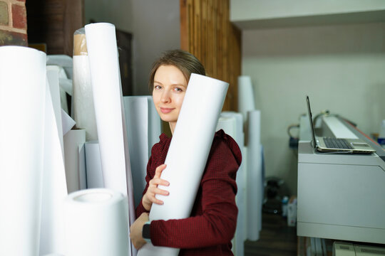 Happy woman holding roll of paper and standing near printer at printing studio