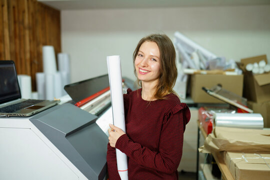 Smiling woman holding roll of paper and standing near printer at printing studio
