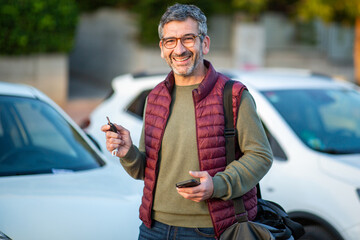 Smiling man holding car key and phone near parked cars