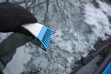 Hand with an ice scraper cleaning the car windshield from frozen snow, important tool for save...