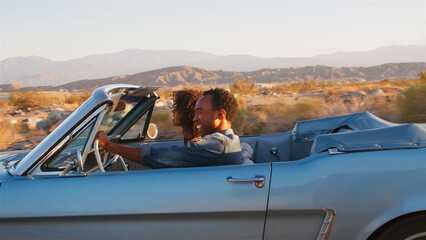 Side View Of Young Couple On Road Trip Driving In Classic Convertible Car Along Country Road