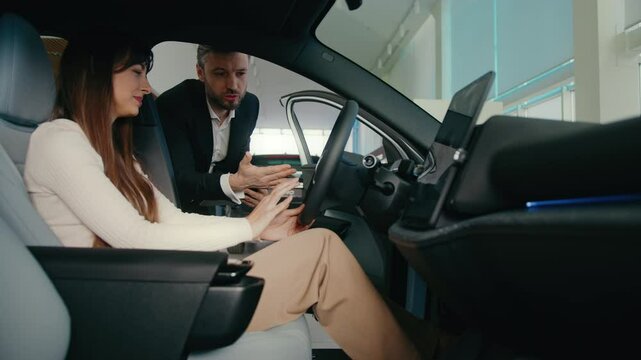 Beautiful Caucasian female sitting inside of new vehicle at drivers seat. Woman carefully listening to dealership manager talking. Man explaining everything about new car. Learning dashboard.