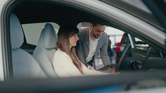 Pretty Caucasian female sitting in car while talking to dealership manager. Asking questions. Man looking at tablet device before answering. Pressing once on button on display of vehicle.