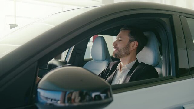 Positive Caucasian male with beard sitting inside of his new car at drivers seat. Inspecting everything in vehicle. Looking at camera while smiling. Getting familiar. Happy about personal success.