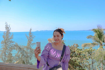 Happy asian woman use smartphone selfie on the sea, tree, blue sky background. Travel Rayong Province, Thailand.