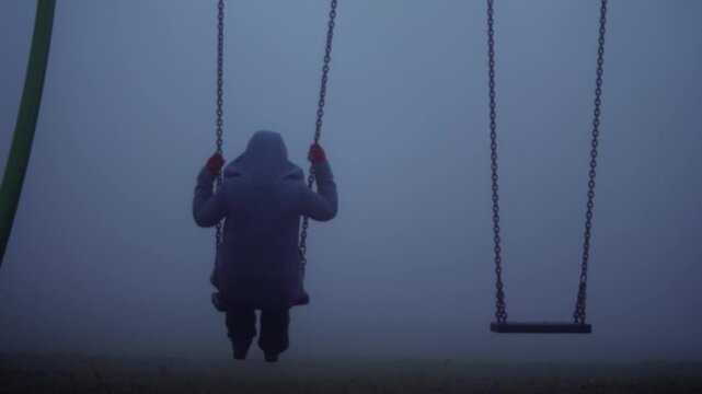 Woman sits on children swings in eerie dark foggy playground