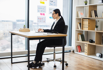 Asian businesswoman in professional attire working at laptop in modern office environment. Young adult focused on tasks, highlighting workplace productivity and focus.