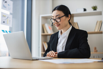 Fototapeta premium Confident Asian businesswoman in headset during video call on laptop. Professional setting, mid-30s, wearing formal suit, focused on business communication, working from modern office with bookshelf.
