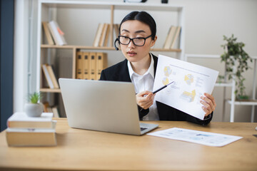 Asian woman in 30s wearing headset engaged in video call, presenting data to colleagues, business setting. Focused and professional atmosphere in modern office space.