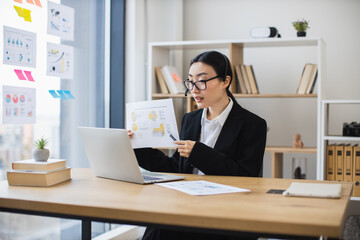 Fototapeta premium Asian female professional in formal attire conducts online meeting, presenting analysis report on laptop. Modern office setting shows productivity, communication, expertise.