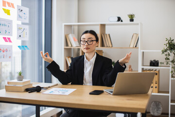 Asian businesswoman meditating at desk, finding balance during office break. Adult female in formal...