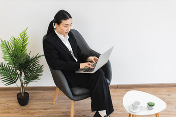 Asian businesswoman in early thirties sitting on chair using laptop in modern office setting with indoor plant. Represents professional work, concentration, technology, business environment.