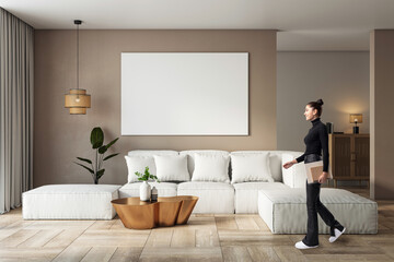 Modern living room interior with beige sofa, blank white canvas on wall, wooden floor, and woman walking by holding a notebook.