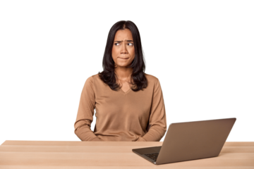 Filipino woman at desk with laptop confused, feels doubtful and unsure.