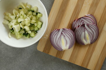 Halved red onion on a cutting board, next to it, diced cucumber and pepper in a white bowl.