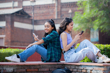 Fototapeta premium Two Modern Friends Using Their Smartphones While Sitting Back to Back