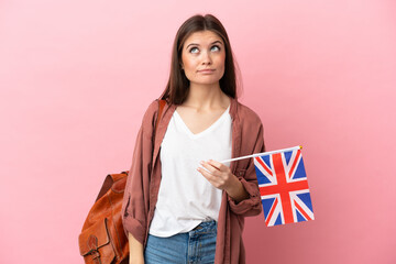 Young caucasian woman holding an United Kingdom flag isolated on pink background and looking up