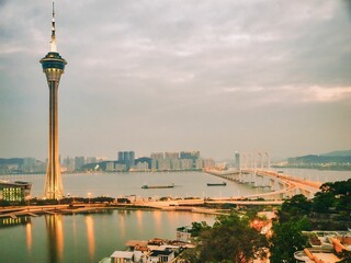 A Scenic View Of Macau City Skyline At Sunset Featuring The Tower And Bridge Illuminated Against A Vibrant Orange And Purple Sky, Reflecting In The Calm Waters Below