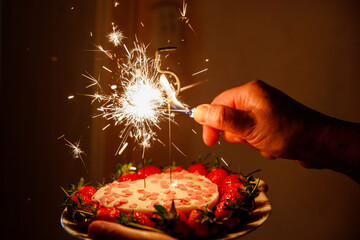 close-up of a heart-shaped cake decorated with fresh strawberries and pink heart-shaped sprinkles. A hand is lighting a sparkler candle placed on top of the cake