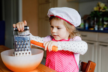 Cute little preschool girl grate carrots for baking carrot cake or cookies. Happy toddler child in...