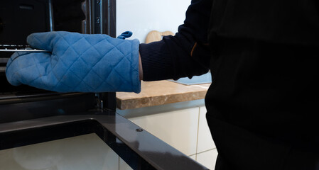 Close up of cheerful man taking baking tray at home with blue oven glove, cooking at home