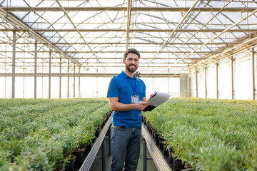 Young bearded man in a blue thisrt standing in a greenhouse and looking enjoyed