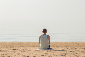 Young adult man sitting alone on sand and looking far away at sea beach in warm sunny summer day....