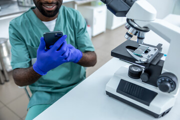 Male chemist using smartphone for online research while analyzing samples under microscope