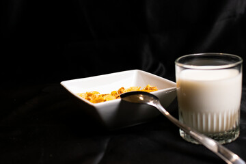 A glass of milk beside a spoon and a bowl of corn flakes cereal. 