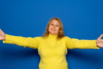 Caucasian woman in her 40s wearing a yellow sweater stretching out her hands, hugging, extending her arms to hug, embracing someone, standing against a blue background