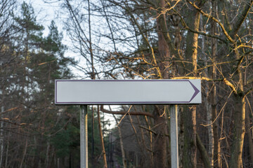 A blank rectangular directional sign with an arrow pointing to the right is mounted on metal posts in a wooded area during daylight; bare tree branches and green pine trees are visible 