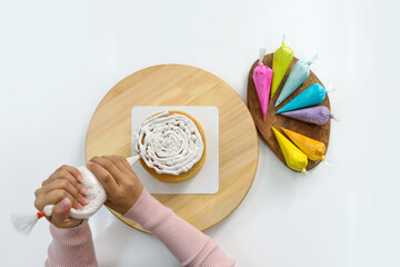 Closeup hand piping white cream on a sponge cake, creating a decorative pattern on the top of a round, light-colored cake that's placed on a wooden turntable or cake stand.