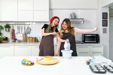 Two women having a great time in a kitchen. Young asian woman in sweater underneath an apron, sing...