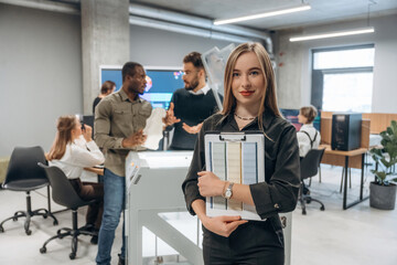 Young woman is standing in front of colleagues. People are in the office by the 3d printer