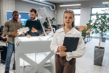 Holding notepad, woman is standing. People are in the office by the 3d printer