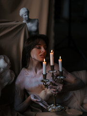 Sexy female sculptor lies in workshop and relaxes after work near antique statues and candlestick with burning candles.
