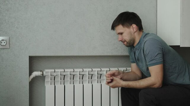 Caucasian man sits near heating radiator and tries to warm up. Portrait of sad person near convector in cold season during breakdown in centralized heating due to energy savings.