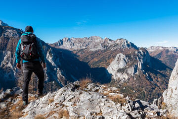 Fototapeta premium Hiker man with backpack looking at mountain peak Griesmauerkogel in Hochschwab Alps, Styria, Austria. Steep rocky cliffs covered in deciduous trees in vibrant autumn colors. Sunny day, Austrian Alps