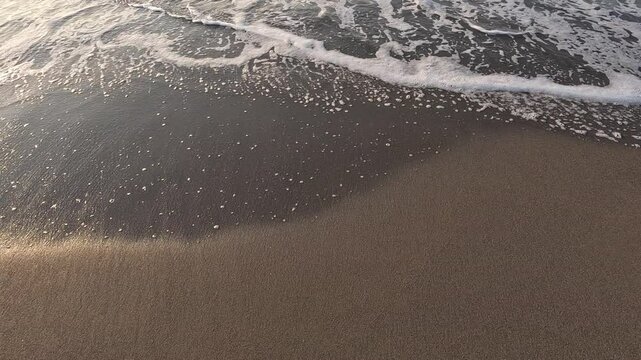 A close-up view of ocean waves gently rolling onto a sandy beach. The foamy white water contrasts with the dark, wet sand as the tide ebbs and flows along the shoreline

