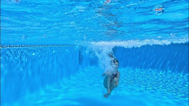A girl jumps doing a cannonball into the swimming pool and dive under the water with a bright, clear blue background