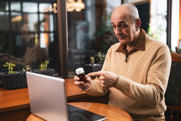 Senior man showing bottle of pills and talking with his doctor during a video call on laptop while sitting in the city cafe.	