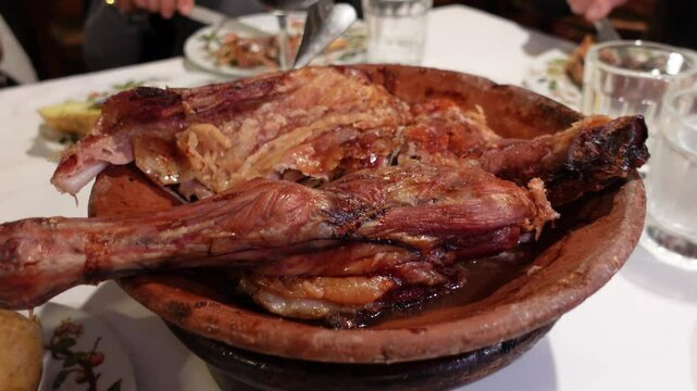 Roasted lamb served in a traditional clay dish with baked potatoes and bread on a dining table, accompanied by red wine and a cozy atmosphere