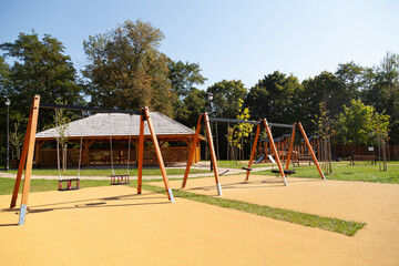 Fototapeta premium Children's playground with swings and a wooden gazebo. Sunny day, lush green trees in background.