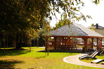 Relaxing wooden gazebo in a park setting, surrounded by lush greenery and a paved pathway. Perfect for a peaceful afternoon.