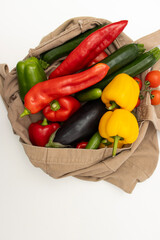 Colorful fresh vegetables including tomatoes, peppers, avocado, eggplant and cucumbers peeking out of brown paper bag on white background. Top view. Healthy eating. Copy space