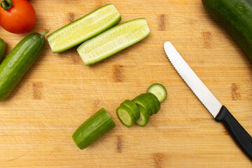Fresh vegetables on a wooden cutting board, including cucumbers, tomatoes, and bell peppers, with a knife for slicing. The arrangement is colorful and neatly presented on a white background with copy 