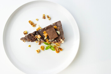 A piece of chocolate cake with nuts, decorated with a mint leaf, lies on a white plate. Small bowls with walnuts and peeled mandarin segments are located nearby. View from above