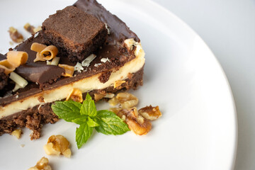 A piece of chocolate cake with nuts, decorated with a mint leaf, lies on a white plate. Small bowls with walnuts and peeled mandarin segments are located nearby. View from above