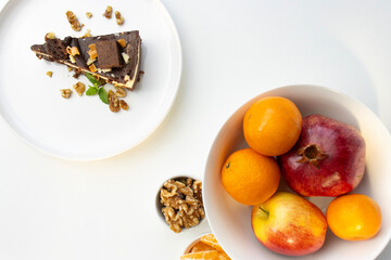 A piece of chocolate cake with nuts, decorated with a mint leaf, lies on a white plate. Small bowls with walnuts and peeled mandarin segments are located nearby. View from above
