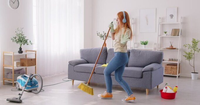 Carefree funny happy young woman, teen girl cleaning house, living room having fun dancing with mop, music in ear headphones overjoyed millennial feels excited to enjoy making chores, sing entertain 